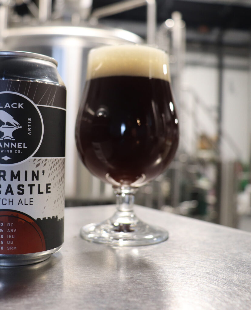Dark Scotch ale with a tan foam head in a stemmed glass beside a Black Flannel Stormin’ the Castle can.