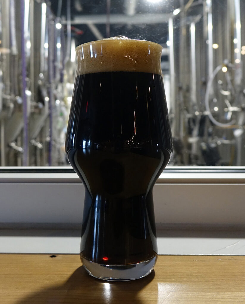 Black American stout with a tan foam head in a curved glass, with brewery tanks in the background.